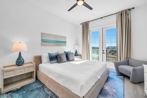 Ein Schlafzimmer mit einem großen Bett, einem Nachttisch, einer Lampe, einem Sessel sowie einem Deckenventilator und Blick ins Freie durch eine Schiebetür.