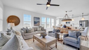 A spacious living room with a sofa, armchairs, and a coffee table. There is a ceiling fan, a chandelier, and a kitchen area in the background.
