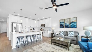 Une cuisine moderne avec des meubles blancs, un îlot central doté de tabourets, un ventilateur de plafond et un coin repas comprenant un canapé et une table basse.