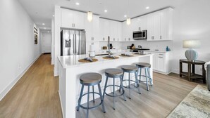 Une cuisine moderne avec des meubles blancs, un îlot central doté de tabourets, des appareils en acier inoxydable et un éclairage suspendu.