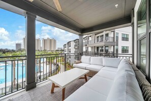 Ein Balkon mit einer weißen Couch, einem kleinen Tisch und Blick auf einen Swimmingpool und Gebäude.