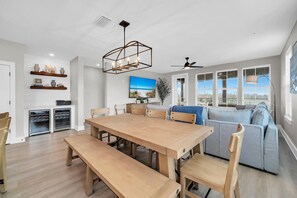 Ein geräumiger Wohnbereich mit Esstisch aus Holz, Bänken, einem grauen Sofa und einem Deckenventilator. Regale mit dekorativen Gegenständen und ein großes Fenster mit Jalousien sind ebenfalls vorhanden.