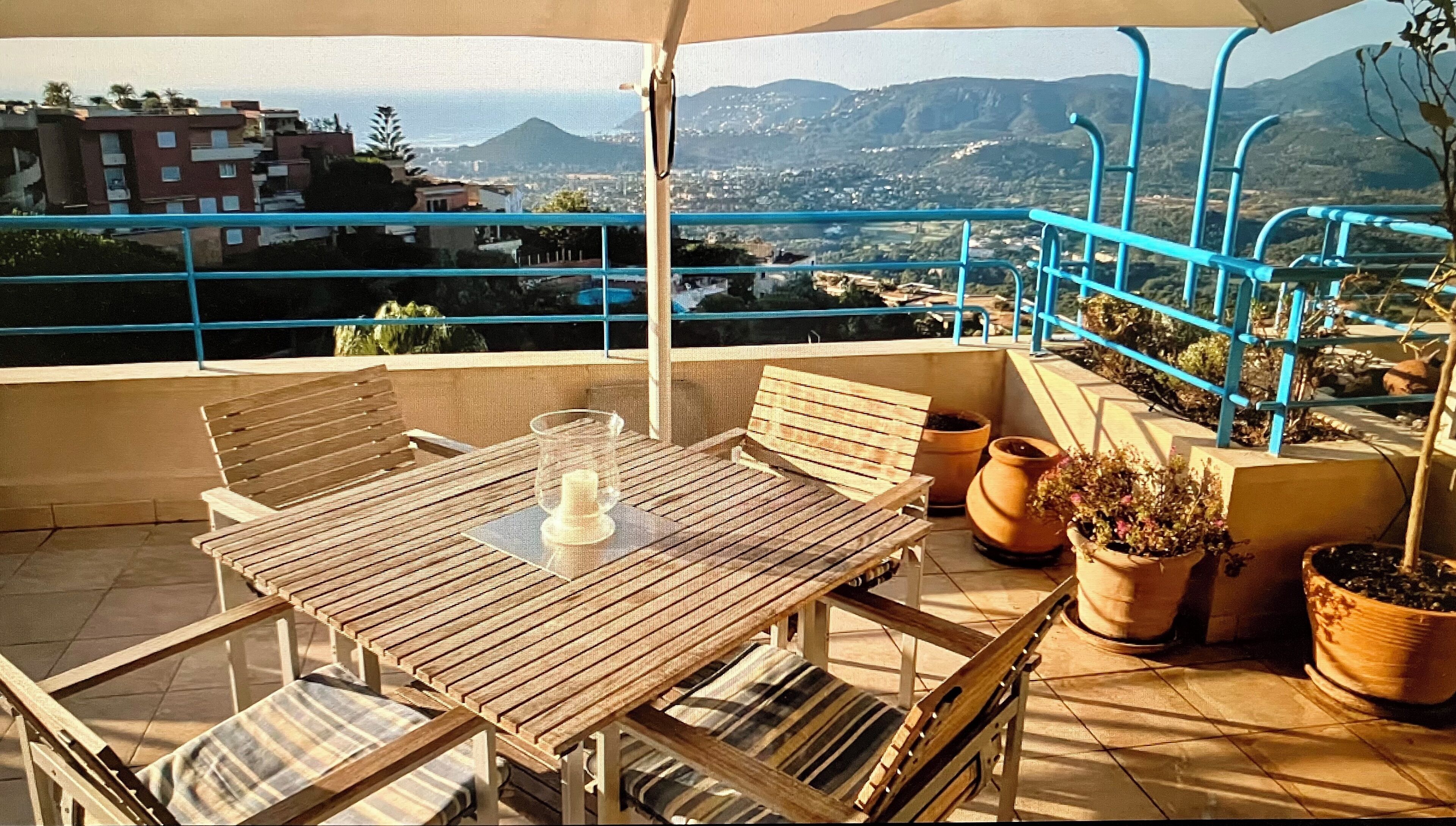 A balcony with a wooden table and chairs, potted plants, and a view of a cityscape and mountains.