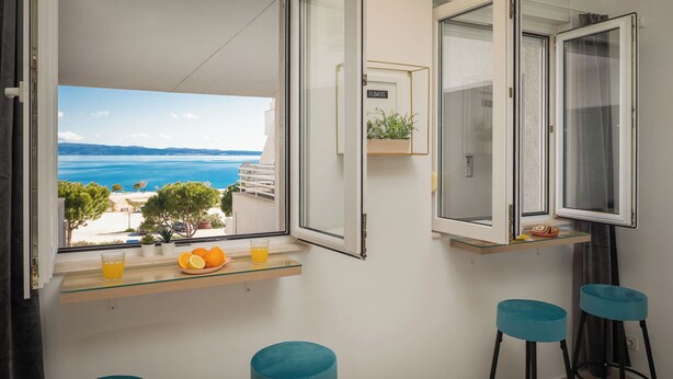 En balkong med utsikt över havet, en glashylla med apelsiner och ett glas apelsinjuice, och två blå barstolar.