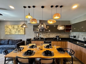 Une cuisine moderne avec un coin repas, comprenant une table en bois dressée pour quatre personnes, un éclairage suspendu et une grande peinture abstraite accrochée au mur.