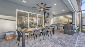Um pátio externo com mesa de jantar, cadeiras, ventilador de teto e área de churrasqueira.