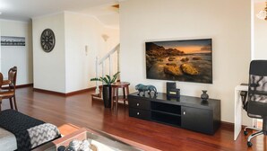 Una sala de estar con piso de madera, una unidad de entretenimiento negra, un televisor de pantalla plana grande, un comedor con mesa y sillas, y una escalera que lleva a otra habitación.