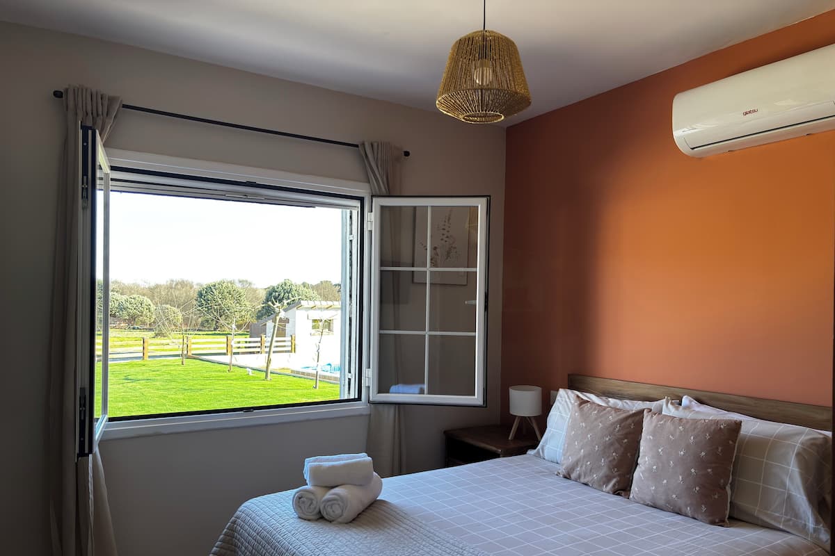 A bedroom with a bed, a window with curtains, a bedside table, and an air conditioning unit.