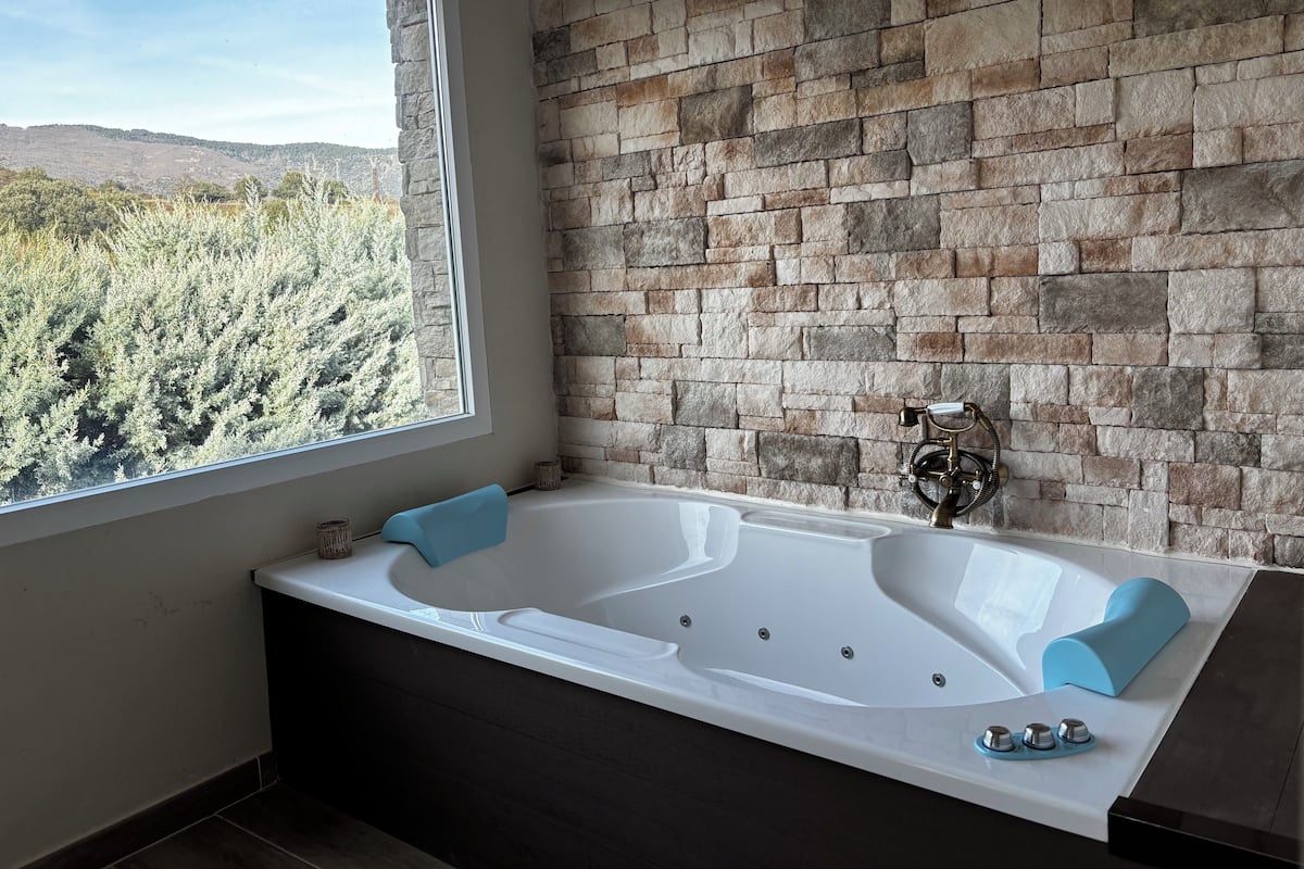 A modern bathroom with a large bathtub, a stone wall, and a view of a forest through a large window.