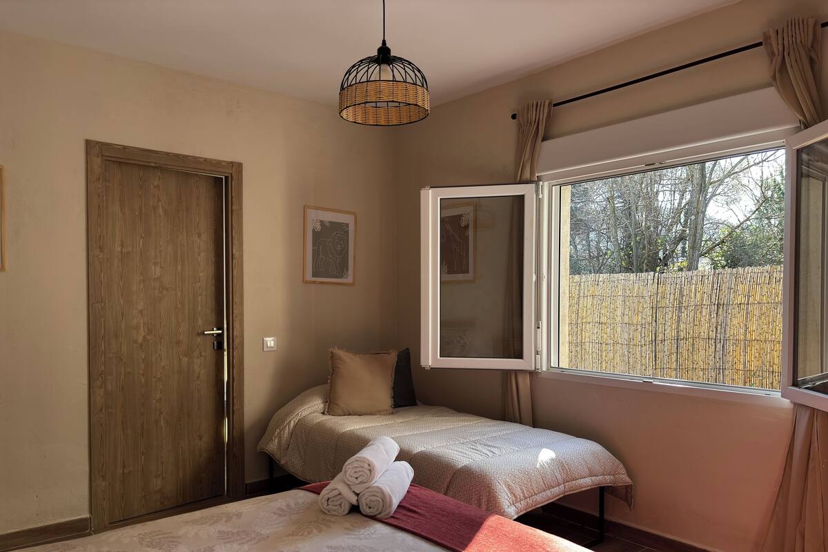 A bedroom with two beds, a wooden wardrobe, a window with a view of a bamboo fence, and a hanging light fixture.