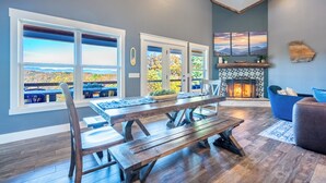 A spacious living room with a large wooden table, a fireplace, and large windows offering a view of the outdoors. - MTN Retreat, 5 Bdrm, Pool, Hot Tub, Game Rm, Theater, Putt-Putt, Fireplace. (Dahlonega)