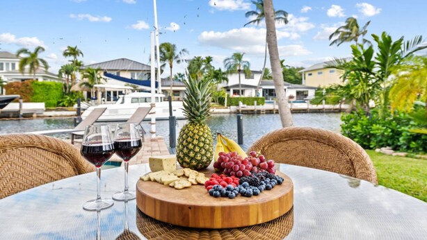 A table with a fruit and cheese board, two glasses of red wine, and a view of a marina with boats and palm trees. - Waterfront Oasis Heated Pool Game Room Gym (Deerfield Beach)