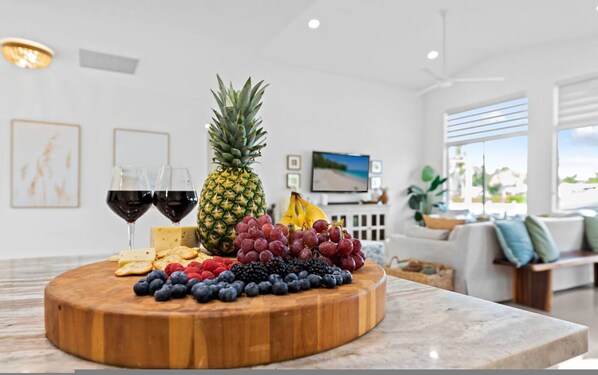 A wooden board with fruits, cheese, and wine glasses on a marble countertop in a modern living room with a TV and plants. - Waterfront Oasis Heated Pool Game Room Gym (Deerfield Beach)