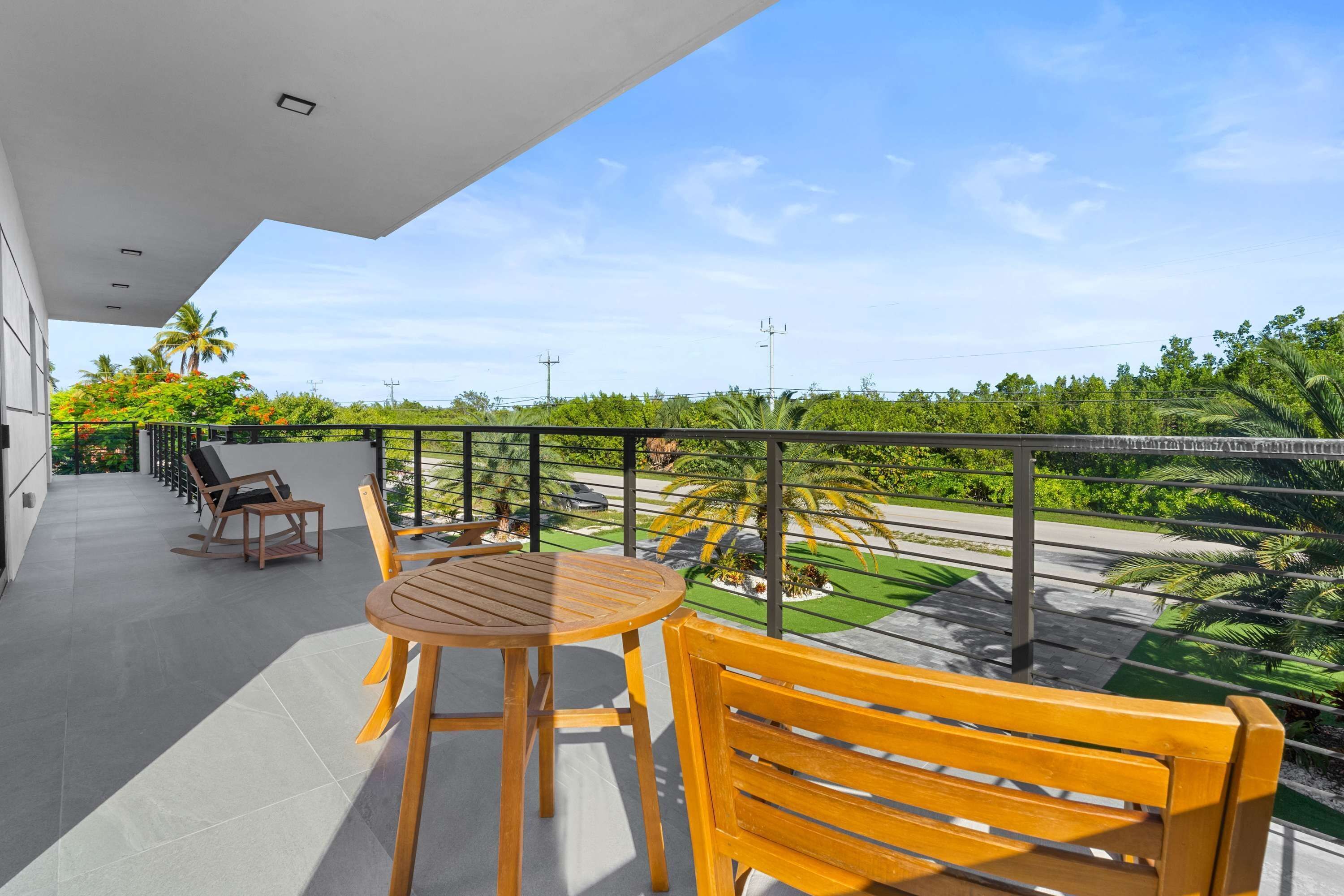 A balcony with a round wooden table and chairs, a wooden bench, and a view of a landscaped area with palm trees and a road.