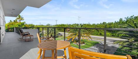 A balcony with a round wooden table and chairs, a wooden bench, and a view of a landscaped area with palm trees and a road.