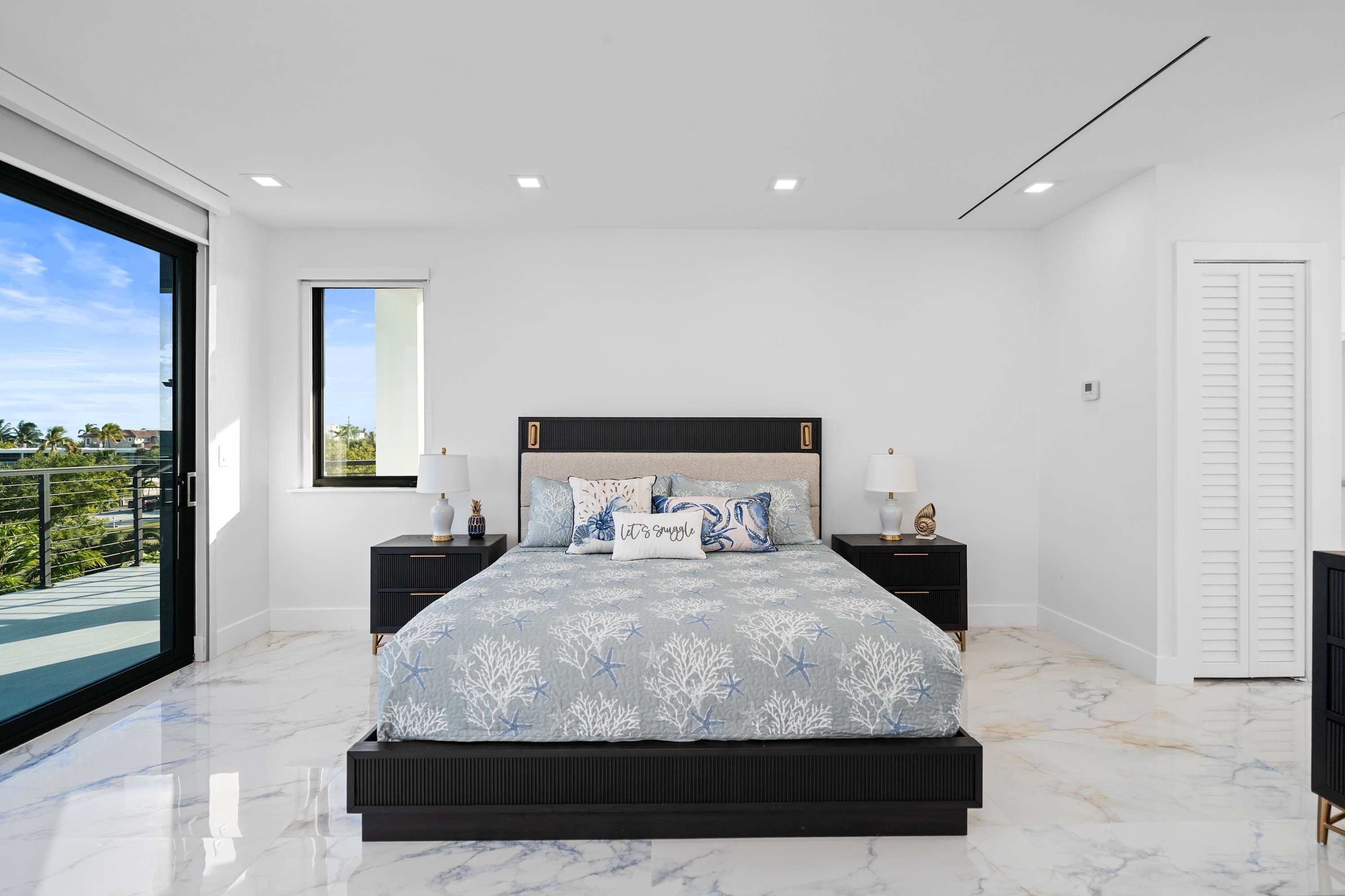 Une chambre moderne avec un grand lit, deux tables de chevet, une vue sur l’extérieur et une porte blanche.