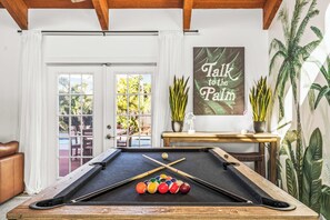 A pool table with balls, a cue stick, and a pool table cloth. There is a door with white curtains, a window with a view of greenery, and a wall with a 'Talk to the Palm' sign.