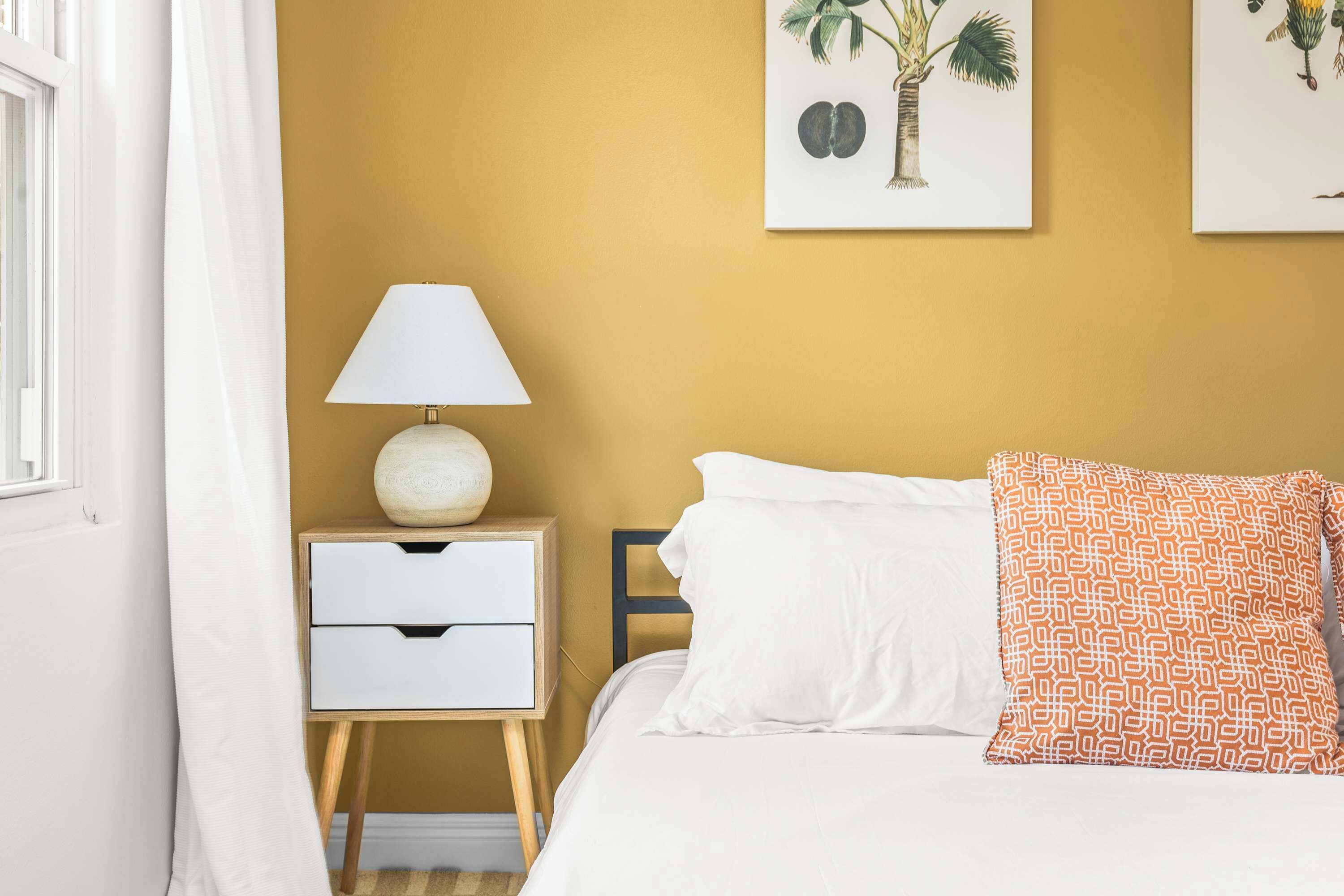 Una camera da letto con un letto, cuscini bianchi, un cuscino decorato, un comodino con una lampada e due opere d'arte a parete.