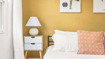 Una camera da letto con un letto, cuscini bianchi, un cuscino decorato, un comodino con una lampada e due opere d'arte a parete.