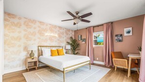 Un dormitorio con cama, ventilador de techo, mesita de noche, silla, escritorio y ventana con cortinas.