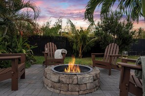A fire pit surrounded by wooden chairs and a bench, set in a garden with a wooden fence and tropical plants.