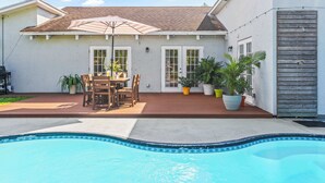 Un patio trasero con piscina, terraza de madera, mesa y sillas de exterior y plantas en macetas.