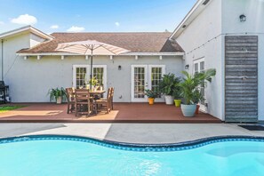 Un patio trasero con piscina, terraza de madera, mesa y sillas de exterior y plantas en macetas.