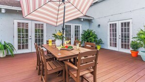 Una terraza de madera con una mesa y sillas para comer, rodeada de macetas con plantas y un paraguas a rayas.