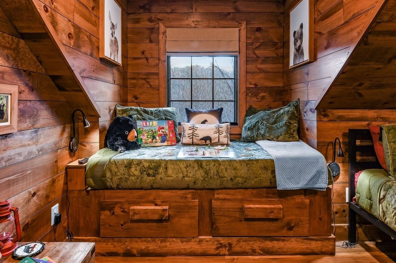 A cozy cabin bedroom with a loft bed, wooden walls, a window with a view, and a small table with a lamp.