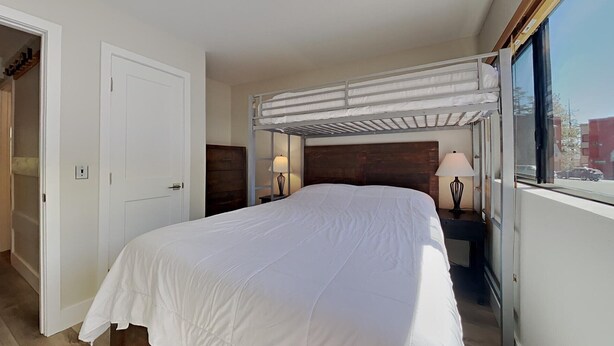 A single-bed room with a bunk bed above, a wooden headboard, bedside lamps, a window with a view of the street, and a door to the left. - Slopeside Condo with Tree View (Mammoth Lakes)