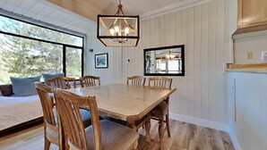 A dining area with a wooden table and chairs, a chandelier, and a large window with a view of trees. - Slopeside Condo with Tree View (Mammoth Lakes)
