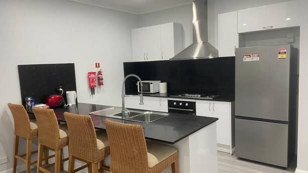 Een moderne keuken met witte kasten, een roestvrijstalen koelkast, een gasfornuis en een magnetron. Drie rieten barstoelen en een zwart aanrecht.