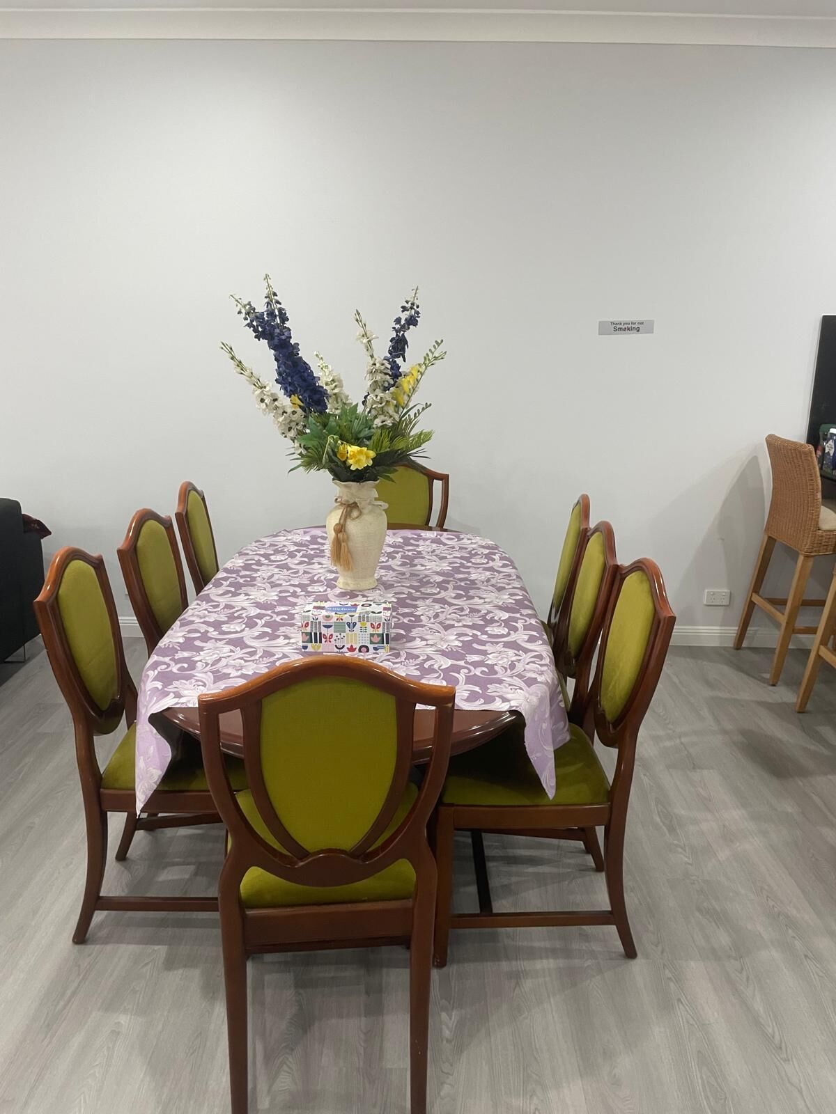 A dining area with a round table covered in a patterned tablecloth, surrounded by green upholstered chairs. A vase with flowers is placed on the table.