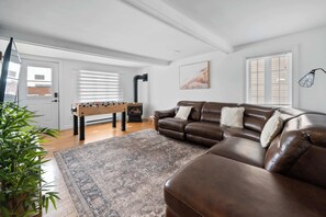 A living room with a brown leather sofa, a wooden coffee table, a foosball table, and a large rug. - SPA | Entre fleuve et montagne | Chalet La Shred (Petite-Rivière-Saint-François)