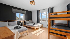 A room with a bunk bed, two single beds, a small table, a lamp, and a window with curtains. - SPA | Entre fleuve et montagne | Chalet La Shred (Petite-Rivière-Saint-François)