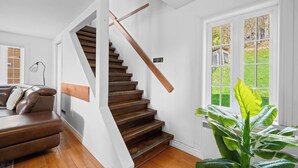 A staircase with wooden steps and a white handrail, a brown leather sofa, a large green plant, and a window with a view of a wooden deck. - SPA | Entre fleuve et montagne | Chalet La Shred (Petite-Rivière-Saint-François)