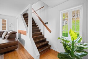 A staircase with wooden steps and a white handrail, a brown leather sofa, a large green plant, and a window with a view of a wooden deck. - SPA | Entre fleuve et montagne | Chalet La Shred (Petite-Rivière-Saint-François)