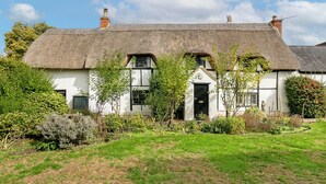 A traditional white house with a thatched roof, surrounded by a well-maintained garden with various shrubs and a lawn. - Lovely 5BD Home with Cinema & Games Room (Marsh Gibbon)