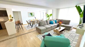 Een moderne woonkamer met een grijze bank, een teal fauteuil en een salontafel. Er is een eethoek met een tafel en stoelen, en een keuken is zichtbaar op de achtergrond.