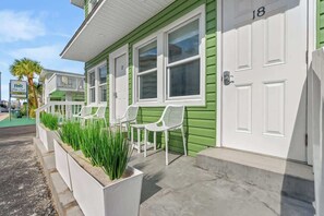 A green and white building with a porch, numbered 18, featuring white chairs and planters with green plants. - Sunny 3BR Near the Beach at Palm Court  (#18) (North Myrtle Beach)