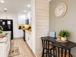A kitchen with white cabinets, a black refrigerator, a wooden floor, a clock on the wall, and a table with chairs. - Captain Chris at South Winds | Across from Beach + Walk to Gulf Shores Fun (Gulf Shores)