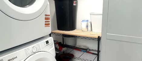 Un espace buanderie avec des lave-linge et des sèche-linge superposés, une poubelle noire, un réfrigérateur blanc et une étagère contenant des produits de nettoyage.