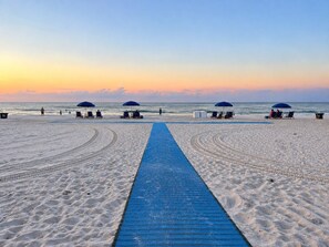 A beach with a blue boardwalk leading to the sea, surrounded by umbrellas and beach chairs. - Captain Chris at South Winds | Across from Beach + Walk to Gulf Shores Fun (Gulf Shores)