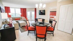 Une chambre d’hôtel moderne dotée d’un coin repas, d’un ensemble de canapés, d’une télévision et d’un balcon avec des rideaux rouges.