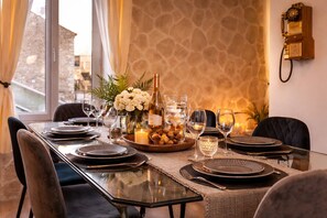 A dining table set with plates, glasses, a candle, and a bottle, with a stone wall and a window in the background.