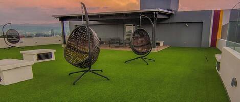 Una terraza en la azotea con césped artificial, dos sillones colgantes y un edificio moderno al fondo.