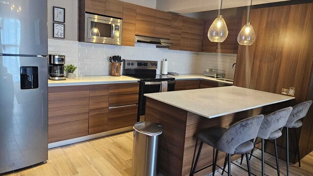 Une cuisine moderne avec des meubles en bois, des appareils en acier inoxydable, un îlot central doté de tabourets de bar et un éclairage suspendu.
