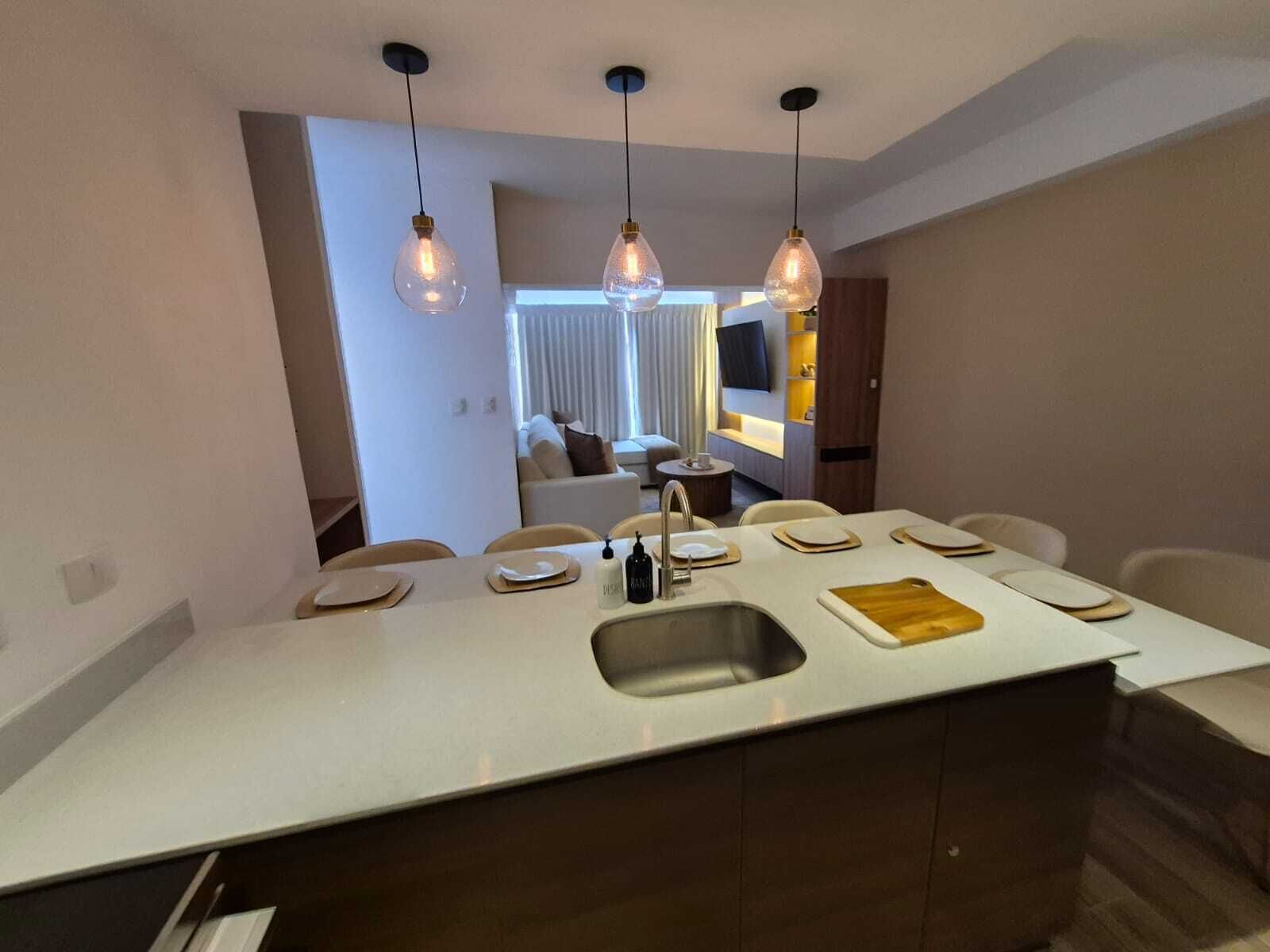 A modern kitchen with a central island, pendant lighting, and a view into a living area.