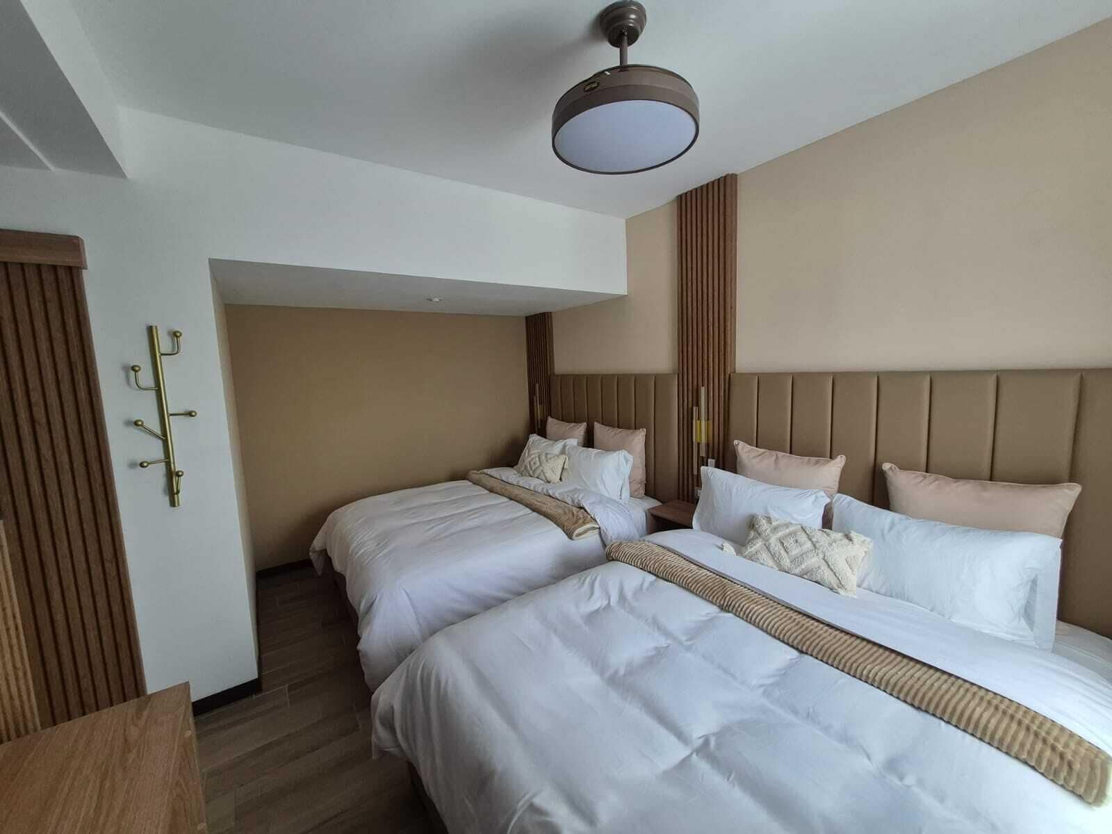 Habitación de hotel con dos camas, cabecera de madera, perchero en la pared y una lámpara en el techo.