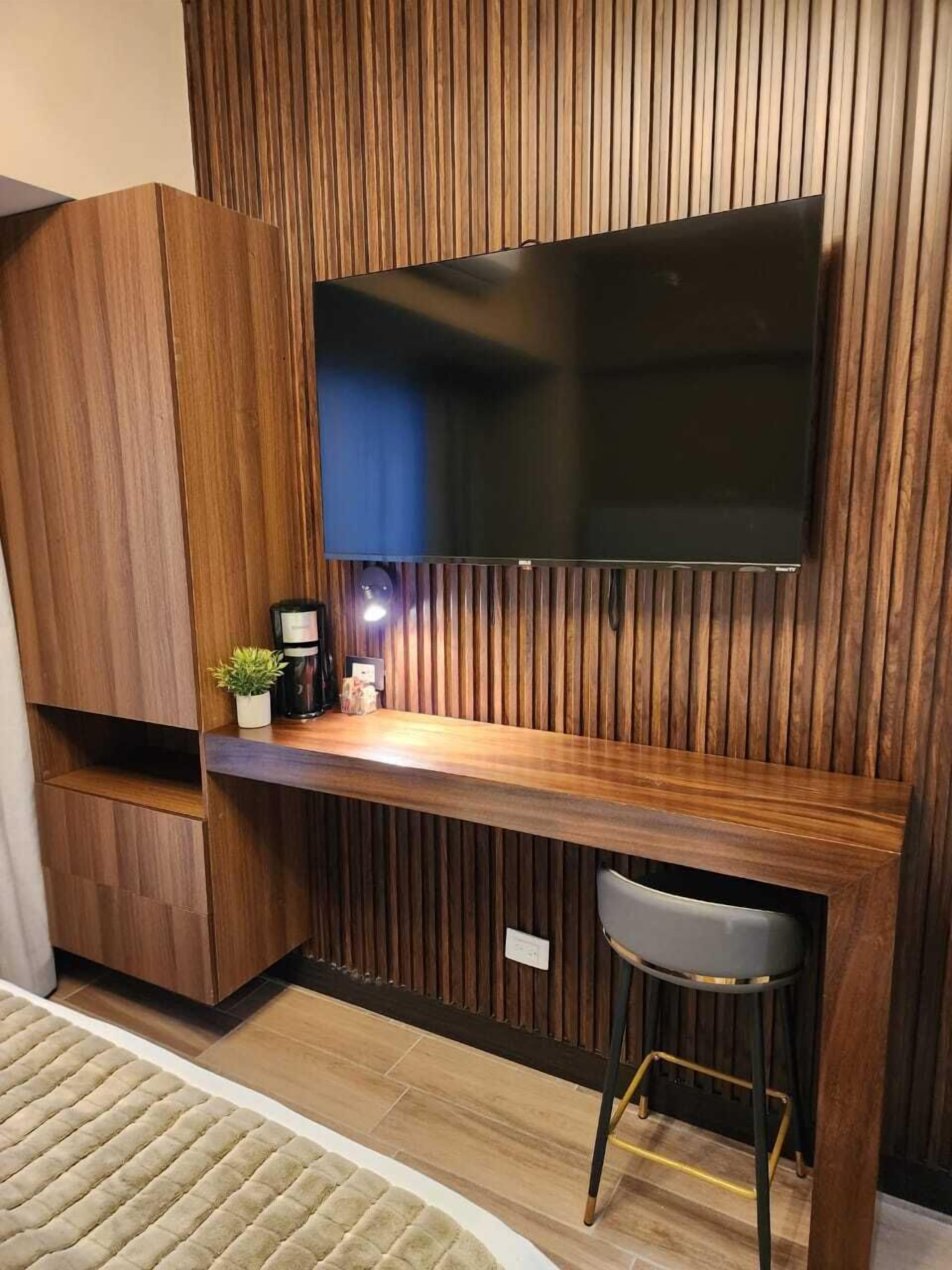 A modern room with a wooden panel wall, a flat-screen TV mounted above a wooden desk, a small stool, and a potted plant.